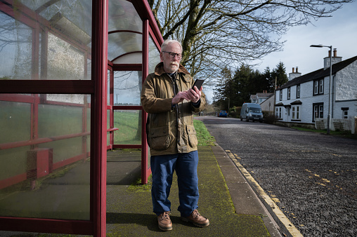 Senior man looking at his mobile phone while standing at a bus stop in a rural location in Scotland