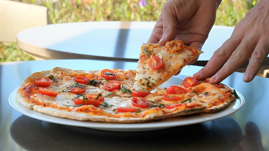 Man hand take a slice of pizza with melted cheese on a plate among other tableware on the table. Fast food