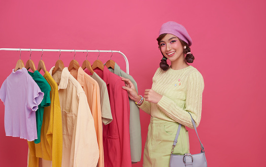 Woman shopping clothes. Beautiful happy smiling asian teen Shopper looking at clothing in store shop isolated on pink background.