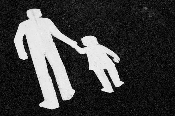 A pedestrian crossing sign consisting of two white figures, an adult and a child holding hands, is painted on an asphalt road surface, indicating a safe area for pedestrians to cross.
