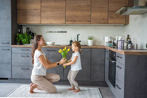 Little girl giving flowers to her mum, while woman cooking on the kitchen. Mothers day, birthday or International Women's Day 8 march concept. High quality photo