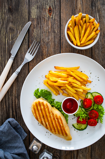Grilled chicken breast with fries and fresh vegetables on wooden background