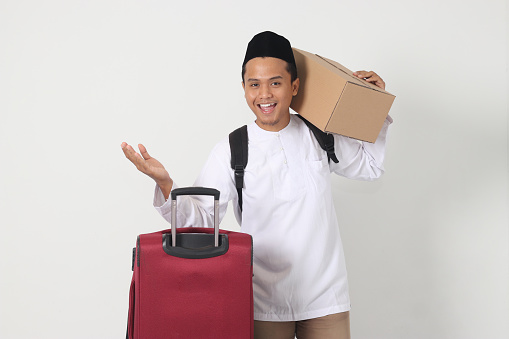 Portrait of excited Asian muslim man carrying cardboard box and suitcase while pointing sideways with finger. Going home for Eid Mubarak. Isolated image on white background