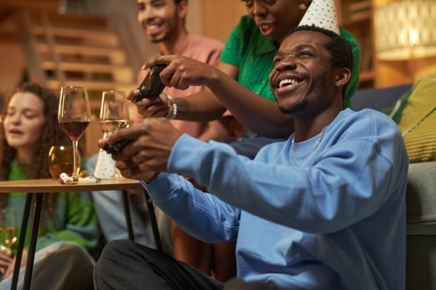 Fun group of adults playing video games during party at home with smiling African American man holding controller