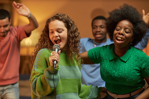 Waist up portrait of carefree young woman singing to microphone and dancing with friends enjoying karaoke together at house party