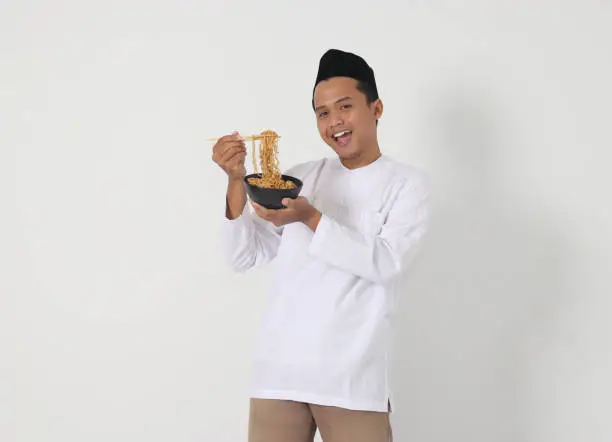 Portrait of attractive Asian muslim man eating tasteful instant noodles with chopsticks served on bowl. Iftar and pre dawn meal concept. Isolated image on white background