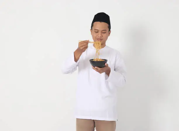 Portrait of attractive Asian muslim man eating tasteful instant noodles with chopsticks served on bowl. Iftar and pre dawn meal concept. Isolated image on white background