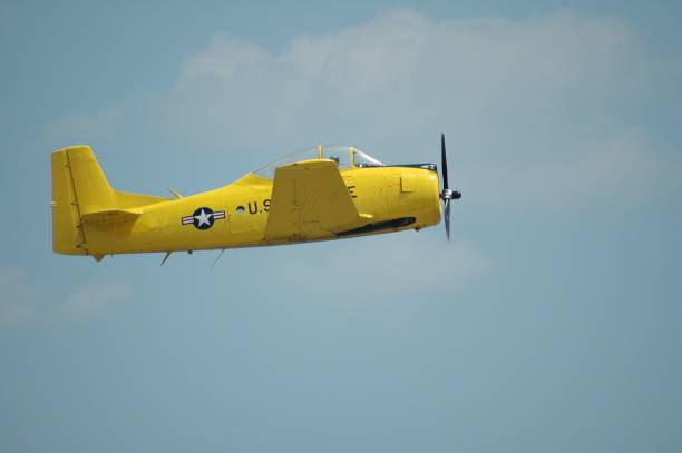 Suwalki, Poland, June 26 2025 (Odlotowe Suwalki 2025) - A Canadian Car and Foundry Harvard Mk.IV, a yellow propeller-driven aircraft, is captured in flight against a cloudy sky. The plane is seen from a head-on perspective, slightly angled, with a visible trail of smoke or exhaust emanating from its rear, indicating it is actively flying, possibly during an airshow or demonstration.