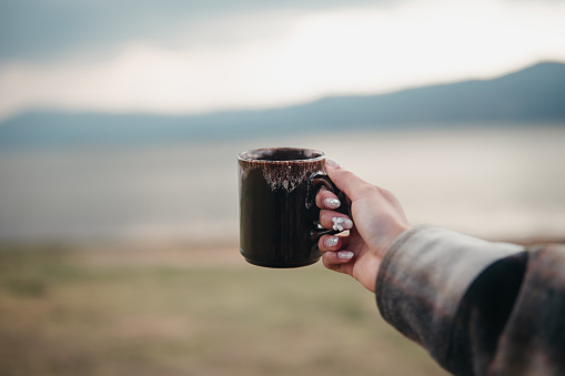 Close-up of a hand holding a dark ceramic mug with a scenic lake and mountain range in the soft-focus background