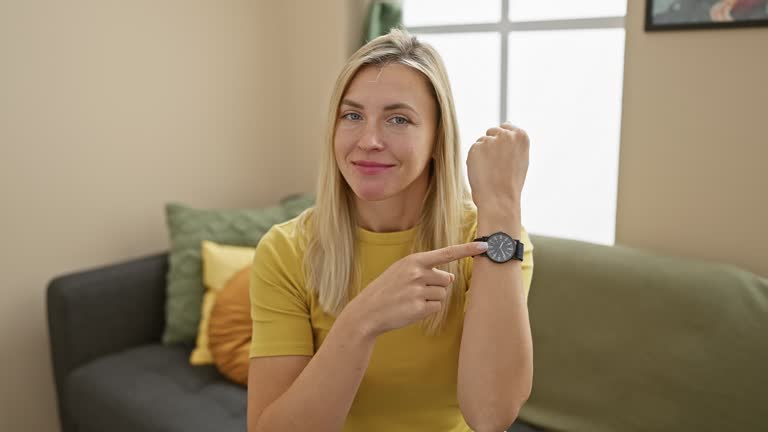 Blonde woman, late for work, pointing at wristwatch in hurry, checks time while casually wearing a t-shirt home