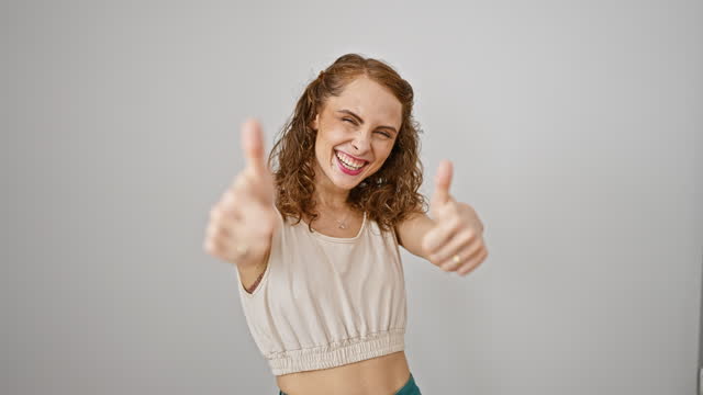 Cheerful young woman gesturing thumbs up, smiling radiantly showing positive winner sign. celebrating success as standing happy against an isolated white background.