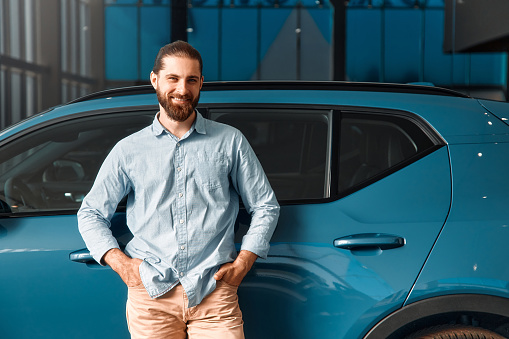 Caucasian bearded man in shirt standing at his dream car in a car dealership. Purchase and rental of cars.
