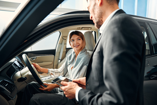 A young woman sitting in a car, examining it in a car dealership and receiving advice from a salesperson with a tablet. Buying, renting and insuring a car.