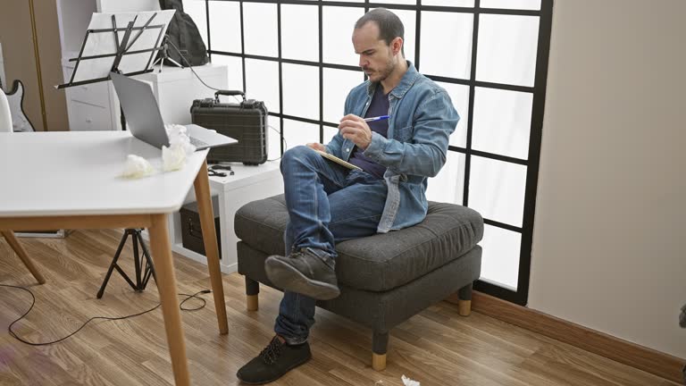 A bald man with a beard writing in a notebook while seated on an ottoman in a studio room with modern decor.