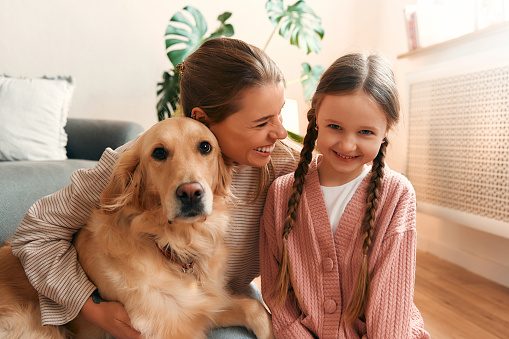 Beautiful mother with cute daughter playing and having fun with dog in cozy living room having a nice time together.