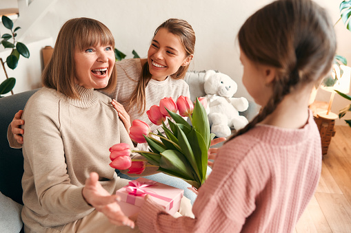 Granddaughter and daughter congratulating their grandmother, giving her flowers and a gift while sitting on the sofa in the living room. Concept of Women's Day and Mother's Day. Women's generation.