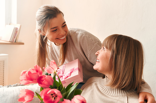 Young woman congratulating her grandmother or mother by giving her flowers and a gift while sitting on the sofa in the living room. Mother's day and women's day concept.