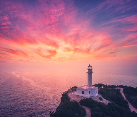 Aerial view of lighthouse on the mountain at colorful sunset in summer. Beautiful lighthouse on the rock, cliffs, sea, sky with pink and purple clouds at dusk. Top view of Cape Lefkada, Greece. Beacon