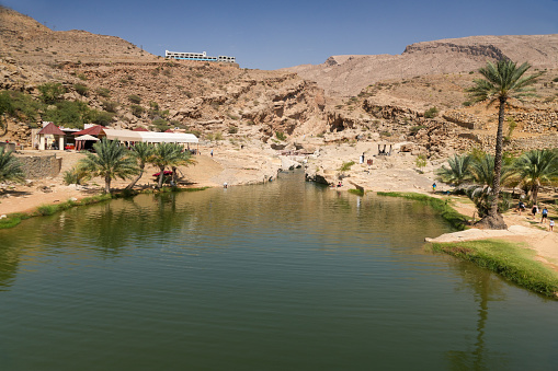 Refreshing water in lake, nice place for swimming in oasis Wadi Bani Khalid, Oman. Palm, small trees, rocks, restaurant and few unrecognized persons walking in background.