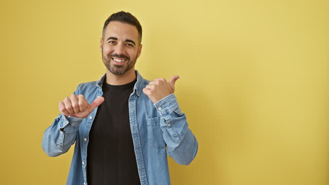 Handsome young hispanic man in denim shirt pointing backwards. confidence written all over his smiling face, thumbs up! on an isolated yellow cutout background