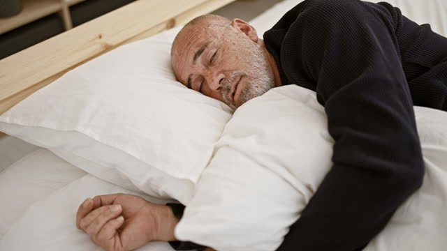 Mature bald man with beard resting in bed, indoors bedroom scene conveying solitude and relaxation, adult male.