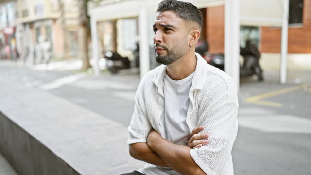 A stressed young bearded man with crossed arms stands thoughtfully on a city street, exuding urban lifestyle and emotions.