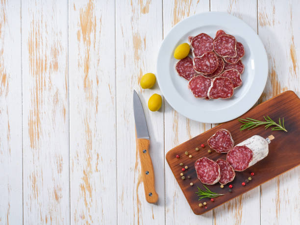 saucisse de fuet coupée en tranches sur une assiette blanche avec des grains de poivre et du romarin frais, sur une table en bois blanc, vue de dessus. - cold cuts thin portion salami photos et images de collection