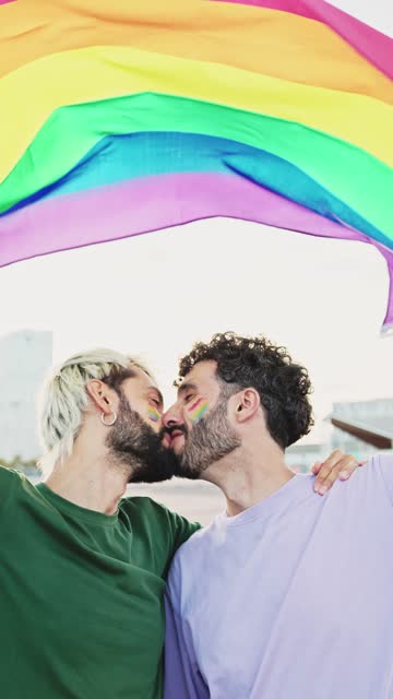 Vertical video of a gay couple kissing in love as they celebrate LGBT Pride Day parade with a rainbow flag