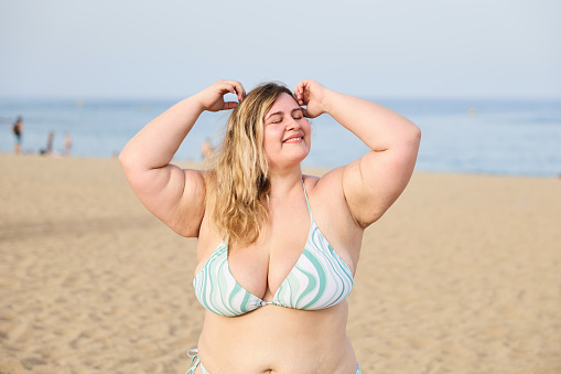 Young smiling plus size female with wavy hair wearing bikini standing on sandy shore against ocean while enjoying summer vacation body positive concept