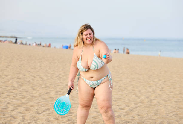 Smiling young overweight female in bikini holding tennis racket and ball while playing on sandy beach near sea. Self-acceptance and body positivity.