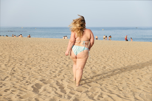 Plus size obese woman in a bikini enjoying a sunny day at the beach on her summer holiday. She is running on the sand.
