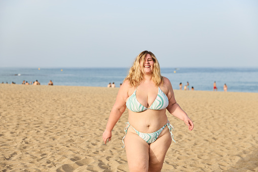 Happy young plus-size woman with wavy hair wearing bikini smiling happily while spending summer holiday on sandy beach against the sea. Concept of a person happy with her body, without complexes.