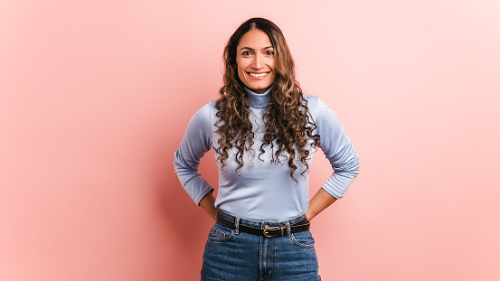 Confident young Colombian lady with long wavy hair in jeans and turtleneck top smiling and standing against pink background in studio while looking at camera