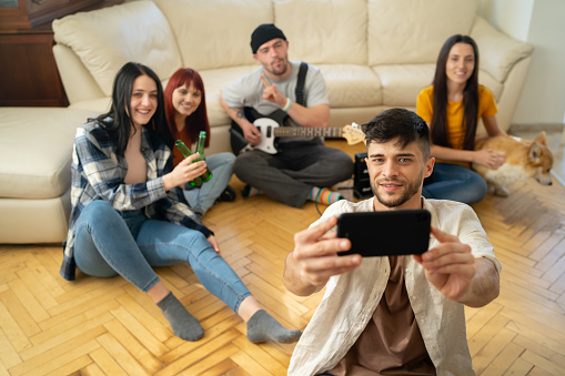 A young man is holding his cell phone to take a selfie of himself and his friends sitting on a living room floor in a modern room