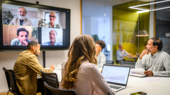 Business people attending a hybrid meeting with some people in a conference room and others attending via video conferencing on a large screen.