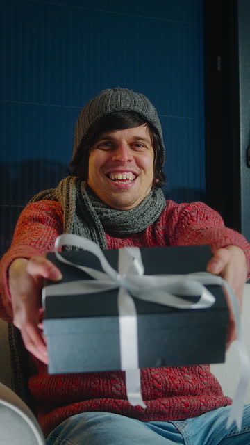 Vertical video. Young man wear scarf and beanie looking at camera and speaking, showing gift box, advertising product, recording video and smiling while sitting on sofa in living room
