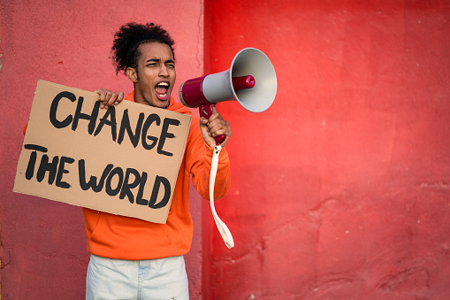 Portrait of furious Afro-American young man wearing an orange sweatshirt, yelling into megaphone. Studio shot on orange background.