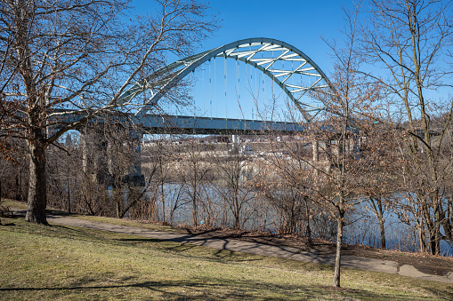 On a sunny spring day, the Birmingham Bridge stretches gracefully over the Allegheny River, framed by a flawless blue sky, as seen from Pittsburgh's Three Rivers Heritage Trail.