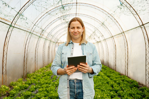 A woman with a pleasant smile stands in the midst of a lush greenhouse, holding a digital tablet that possibly contains records or controls for the greenhouse operations.