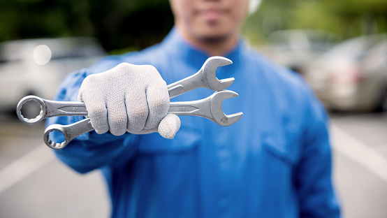 Close-up of professional car mechanic's hands, wearing a blue work uniform and holding a large wrench, as he works on a vehicle. Midsection shot.