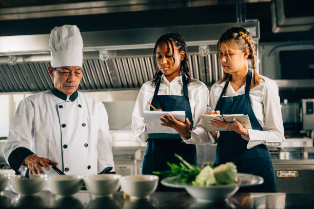 istock Professional Asian senior chef teaching cooking techniques to diverse students in a restaurant kitchen. Focus on teamwork learning note-taking in this educational workshop setting. 2057147518