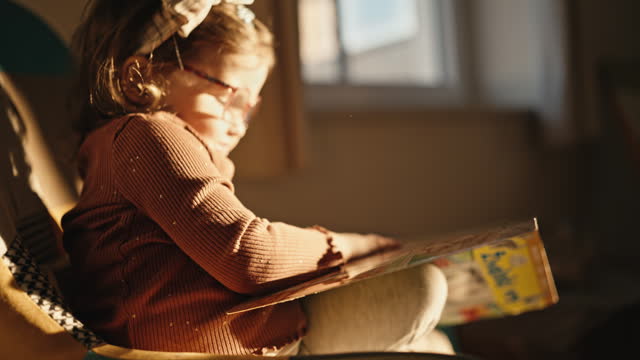 SLO MO Side View of Cute Little Girl Reading Picture Book Sitting on Chair at Home