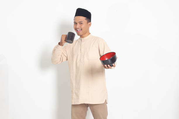 Portrait of excited Asian muslim man in koko shirt with skullcap holding a mug and empty bowl. Bowl template for food brand. Isolated image on white background stock photo