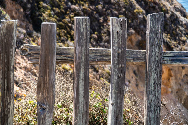 A rustic and aged wooden fence with weathered, pointed planks stands against the backdrop of a verdant landscape in Mallorca. The serene countryside and blue sky evoke a tranquil atmosphere.