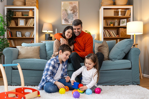 Cute kids playing while parents relaxing sofa at home together, smiling active boy entertaining with toy car near his sister on floor, happy family spending time together in living room on weekend