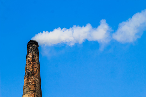 The chimney of brick field emitting heavy smoke with unhealthy carbon di oxide responsible for damaging ozone layer. Factory smoke causes pollution in the air. Fuming chimney against blue sky.