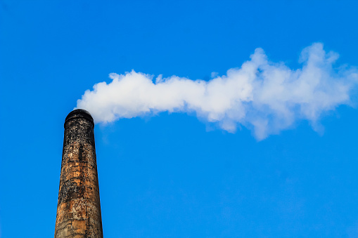 The chimney of brick field emitting heavy smoke with unhealthy carbon di oxide responsible for damaging ozone layer. Factory smoke causes pollution in the air. Fuming chimney against blue sky.