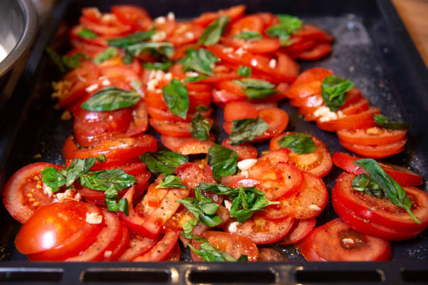 Ripe tomato slices seasoned with garlic and pepper, garnished with fresh basil leaves on a baking tray, ready for roasting.