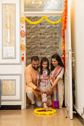 Happy indian family of three doing grah pravesh ritual or entering new house for the first time. daughter entering house with right feet stepping in a plate filled liquid kumkum. Griha Pravesh Concept