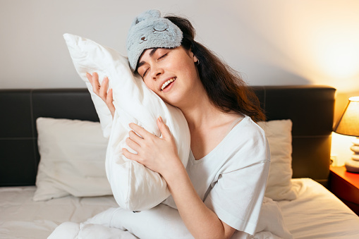 Portrait of pretty smiling woman wearing sleep mask hugs pillow while sitting in bed. Cozy bedroom. Concept of deep sleep and insomnia.